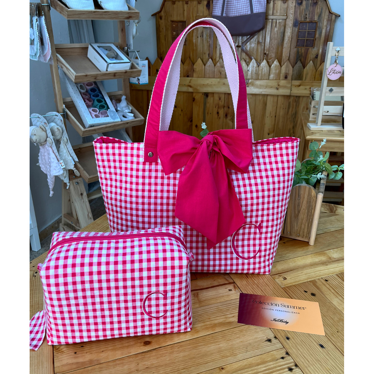 Primer plano de un bolso de playa artesanal de vichy fucsia y neceser personalizado sobre mesa de madera en taller de costura con tarjeta de colección.
