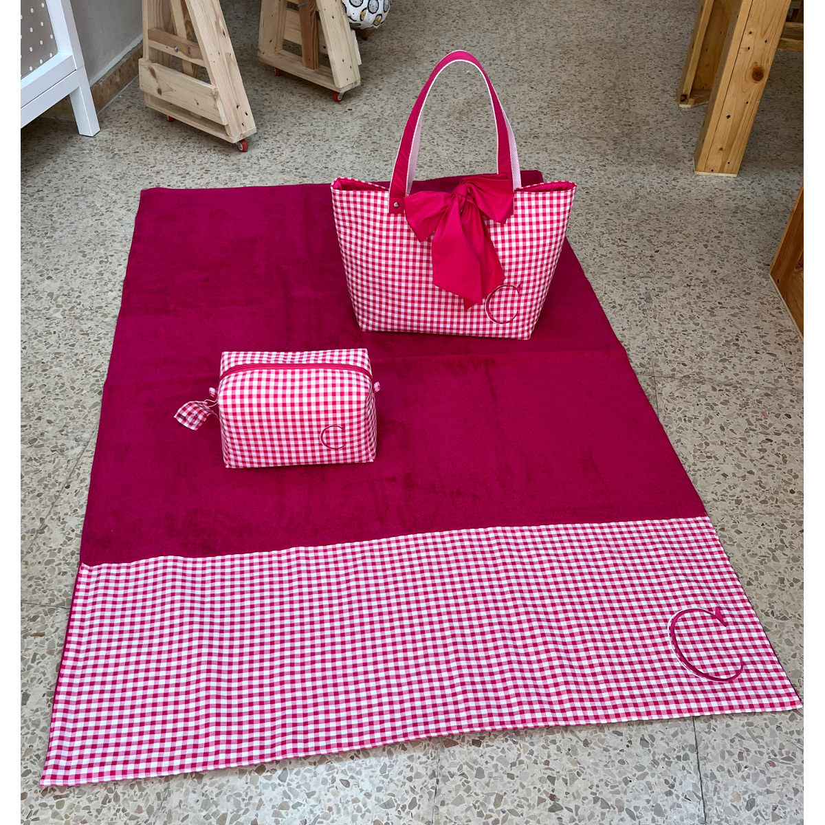 Toalla de playa fucsia abierta con ribete de cuadros vichy junto a un bolso playero y un neceser a juego con la inicial C bordada sobre suelo exterior.