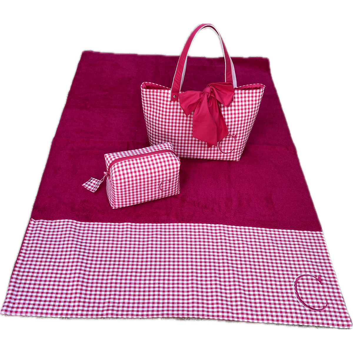 Set de piscina personalizado sobre fondo blanco con toalla de rizo fucsia, neceser de cuadros y bolso de playa con lazo y la inicial C bordada.